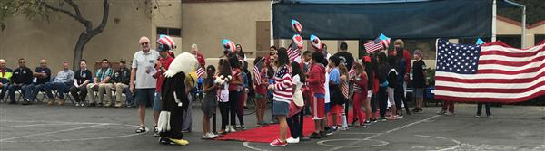 Photo of several military veterans sitting, watching as a veteran is honored near a red carpet lined by students.